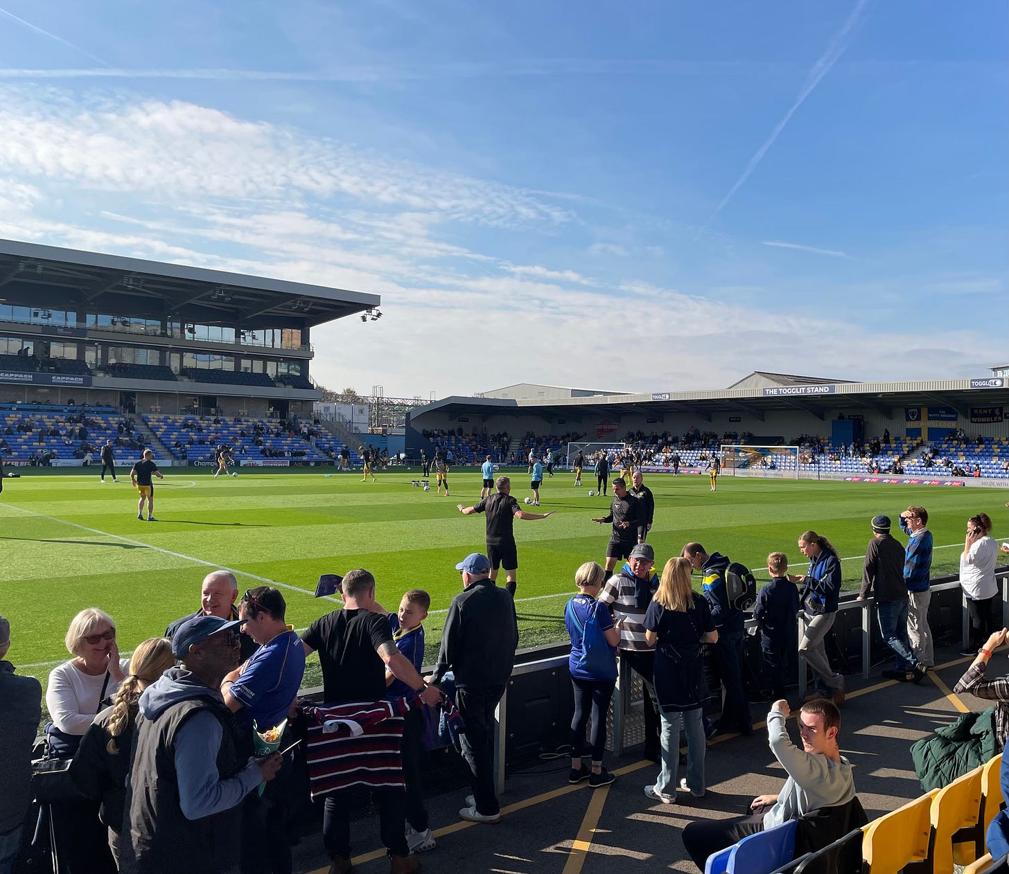 The pitch at Plough Lane football ground in the sunshine with the teams warming up in the background.