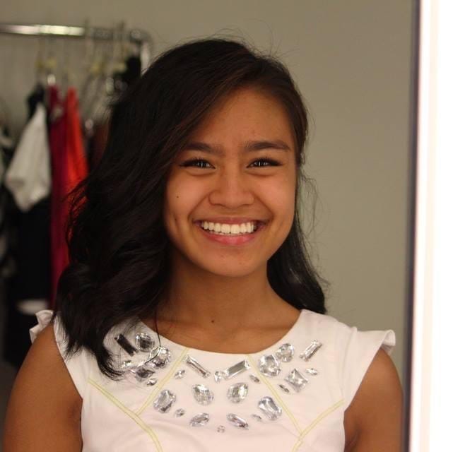 Joelle smiling into the camera through her reflection in the mirror. The left side of her hair is curled while the right side is pinned back behind her shoulder. She wears a white dress wih silver crystals along the neckline