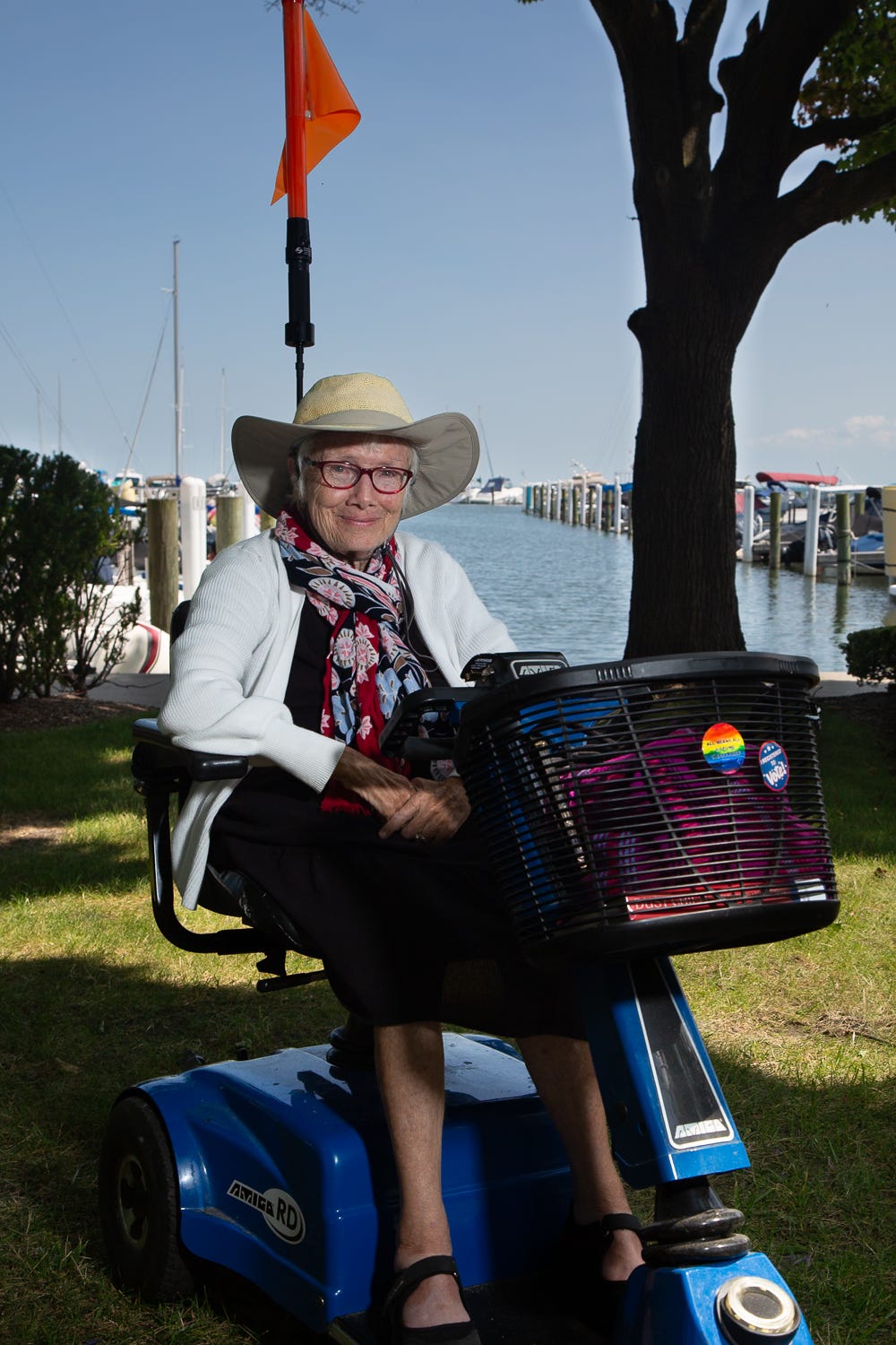 A close portrait of Patricia Lay-Dorsey sitting on a blue mobility scooter at a marina. She smiles at the camera while wearing a wide-brimmed hat, glasses, a patterned scarf, and a white cardigan, with boats and water in the background. A close portrait of Patricia Lay-Dorsey sitting on a blue mobility scooter at a marina. She smiles at the camera while wearing a wide-brimmed hat, glasses, a patterned scarf, and a white cardigan, with boats and water in the background.
