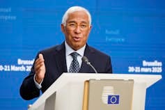 A tanned man with white hair speaks at a podium. A tanned man with white hair speaks at a podium.
