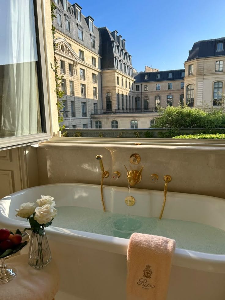 This may contain: a bath tub sitting in front of a window next to a vase with flowers on it