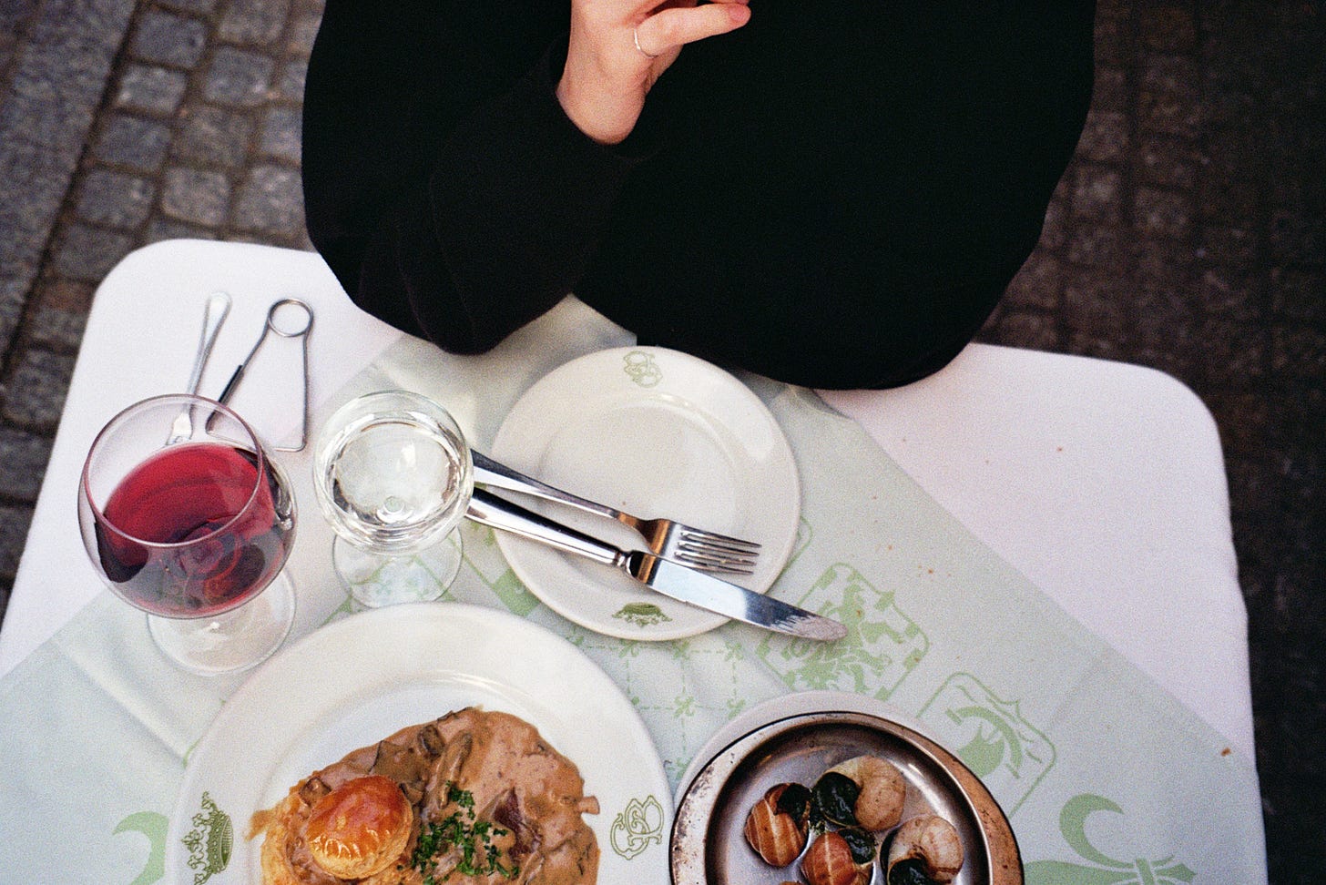 A bird’s eye view of Evyn Silver’s elbows resting on a table on a cobblestone street. The table is covered in white and green table cloths and includes a glass of red wine, escargot, and mushroom vol au vent.