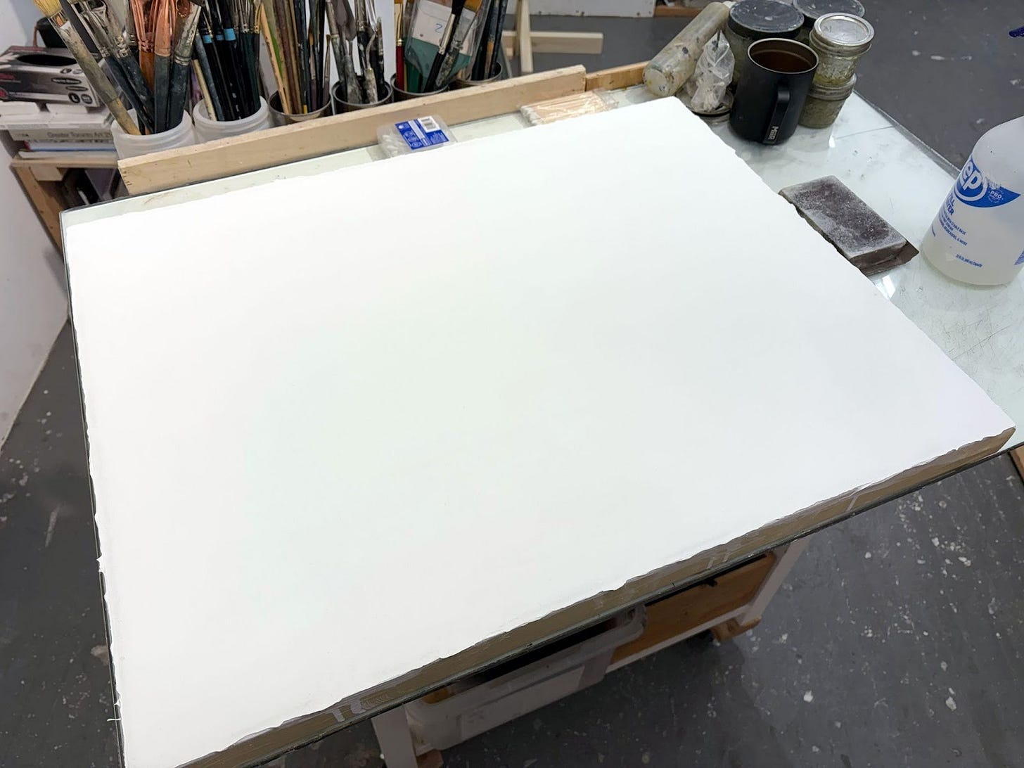 A completed white-tinted molding paste ground on canvas sits atop a studio table surrounded by brushes and other painting tools. The surface of the painting ground is extremely smooth and white.