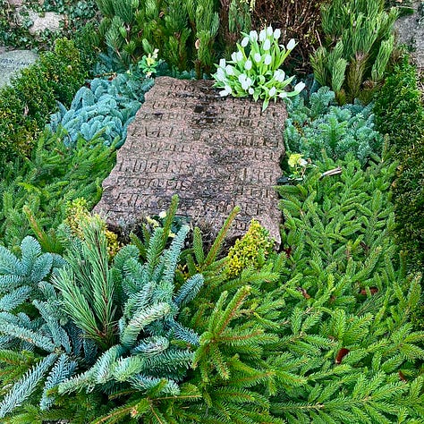 Gravestone with branches of different colored evergreens; neat plots linedwith gravel and divided by small hedges; a granite cross etched with many names