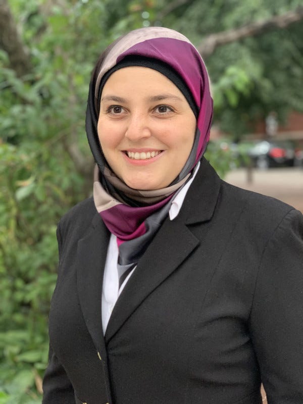 Mona Minkara smiling broadly and looking beyond the camera in front of a pine tree. Dressed in a multiply colored hijab in purple, gray and black that flows into a white open-collared shirt as a scarf. Mona is in a black buttoned-up blazer.