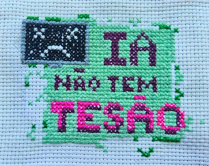 bordado em ponto cruz em que se lê em letras garrafais rosas e roxas: IA NÃO TEM TESÃO. Há um monitor de computador com tela escura e dois olhos como X e uma boca virada pra baixo. O fundo é de um verde menta, que se pixela nas bordas bordado em ponto cruz em que se lê em letras garrafais rosas e roxas: IA NÃO TEM TESÃO. Há um monitor de computador com tela escura e dois olhos como X e uma boca virada pra baixo. O fundo é de um verde menta, que se pixela nas bordas