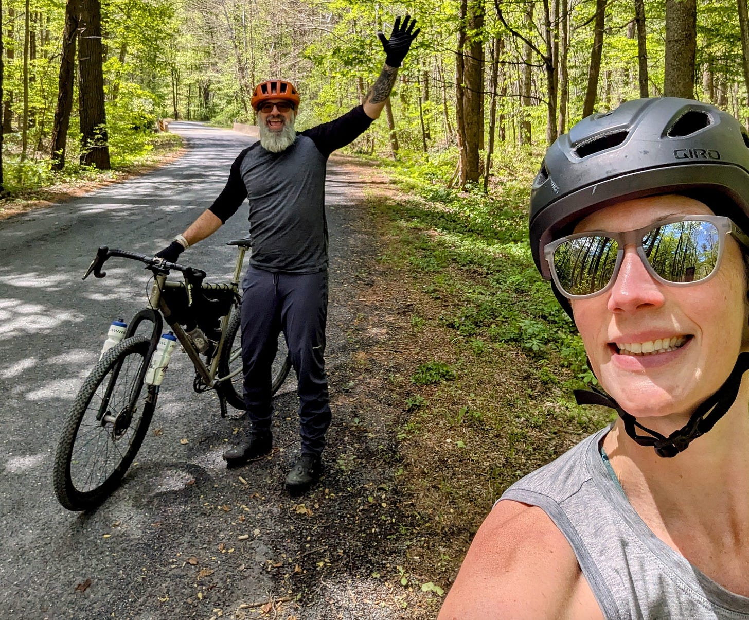 The author and his wife, wearing bike helmets, take a selfie on a quiet forest road in the George Washington National Forest. The author stands in the background beside a gravel bike, waving, while his wife smiles in the foreground. Tall green trees line both sides of the road under a bright blue sky. The author and his wife, wearing bike helmets, take a selfie on a quiet forest road in the George Washington National Forest. The author stands in the background beside a gravel bike, waving, while his wife smiles in the foreground. Tall green trees line both sides of the road under a bright blue sky.