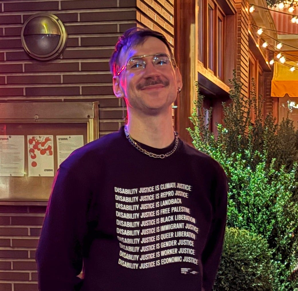 A white disabled queer with a mustache is smiling at the camera outside a restaurant. They are wearing a black sweater with wavy white text. Each line is a sentence that starts with “Disability Justice is…” Climate Justice, Repro Justice, Landback, Free Palestine, Black Liberation, Immigrant Justice, Queer Liberation, Gender Justice, Worker Justice, Economic Justice.