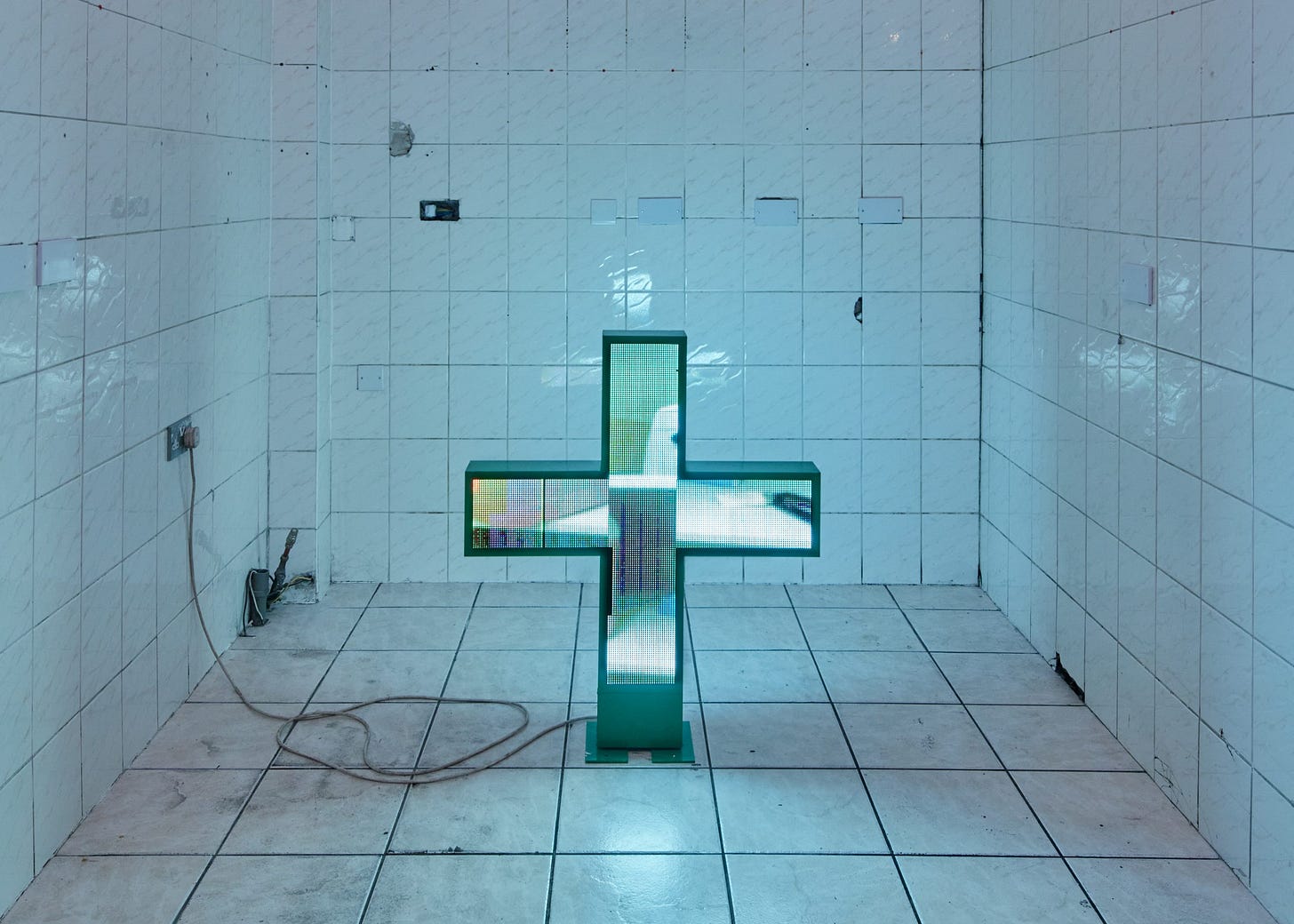 An LED pharmacy sign glows in a white tiled space. It looks like a cross. The LED lights show an indistinct image. The space is grimy. A power cable is visible.