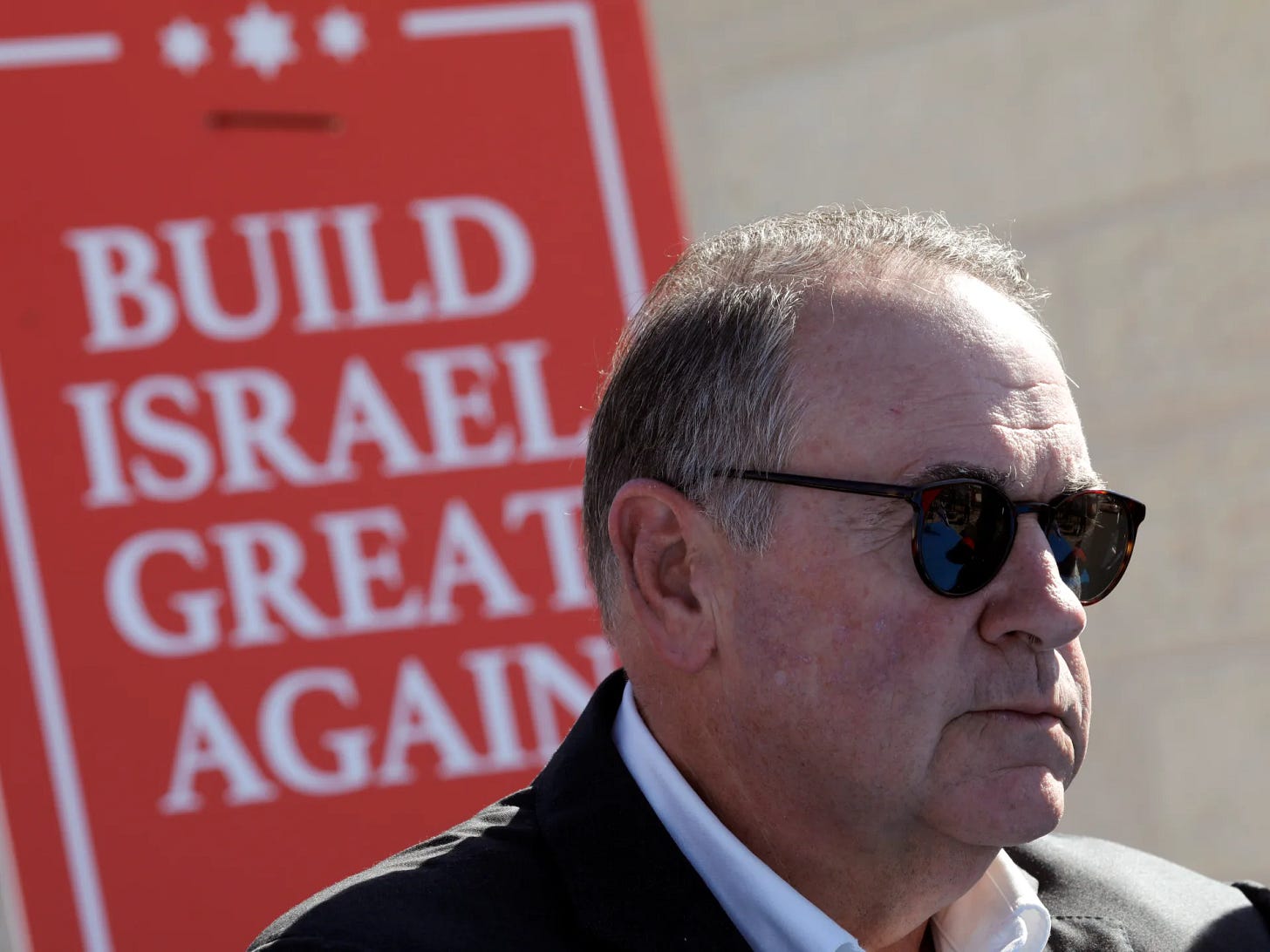 Mike Huckabee, con gafas de sol y una chaqueta oscura, fotografiado de perfil ante un gran cartel rojo con el lema «Build Israel Great Again», variante local del MAGA (Make America Great Again), acuñada en 2018 por los colonos «oficiales» del Yesha Council (Consejo de «Judea y Samaria»).