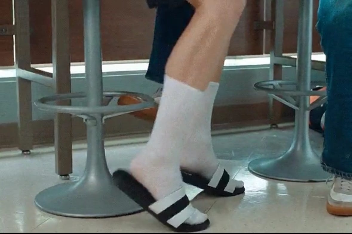 Still from the film Challengers showing the bottom half of two mens' legs in front of stools. One of the men is wearing crew socks and slide sandals.