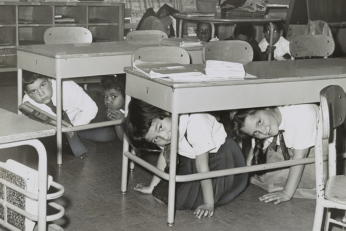 This historical black-and-white photograph from 1962 shows students at P.S. 58 in Brooklyn, New York, practicing a Cold War "duck and cover" air raid drill by crawling under their school desks.