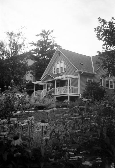 A black and white photograph of a two-storey house surrounded by trees and flowers