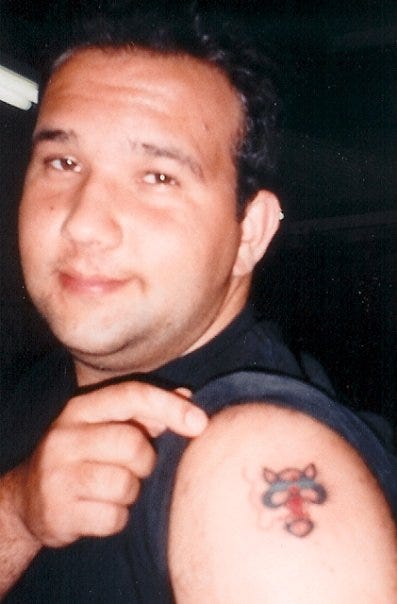 This is a picture of Mickey Fisher at age 21, looking into the camera as he rolls up his left sleeve to show a fresh tattoo on his shoulder. It's the logo for the band Blues Traveler, a cat with sunglasses and a joint hanging out of its mouth.
