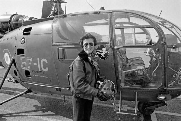 A black-and-white photo of Dr. Valérie André, wearing aviator sunglasses, a leather bomber jacket and holding a helmet in her hands, poses in front of a military helicopter.