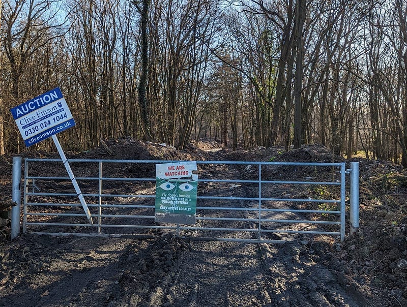 Entrance to a woodland with muddy track, wide metal gate with 2 signs. One Cliver Emson auctioneers and the other We are watching, No Flytipping.