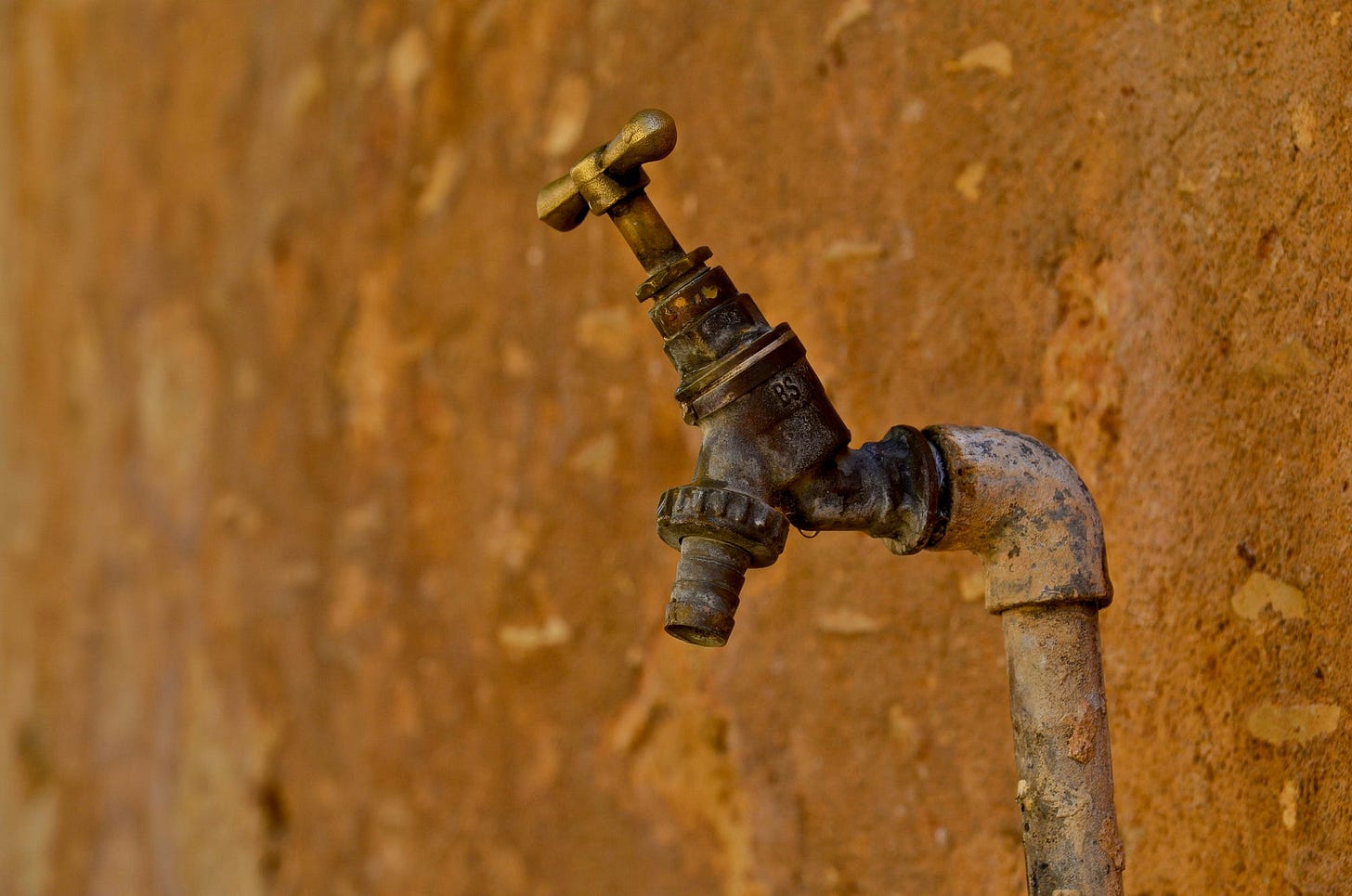 Dry and rusted water spigot next to a crumbling clay building.