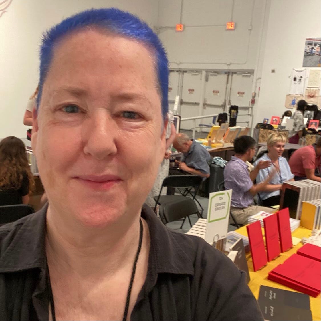 A selfie photo of Sal Randolph, a white woman with very short deep blue hair, in front of the dispersed holdings book table at the Printed Matter Los Angeles Art Book Fair in 2023. In the background you can see black and red dispersed holdings books displayed against a sunflower yellow cloth. Other publishers and buyers are talking with one another and perusing available books. 