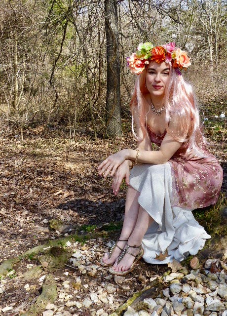A rose-haired, flower-crowned Persephone sits on a mossy rock in a forest just coming into bloom, ankles and wrists crossed, wearing a secretive smile as she dreams of the god who has stolen her heart.