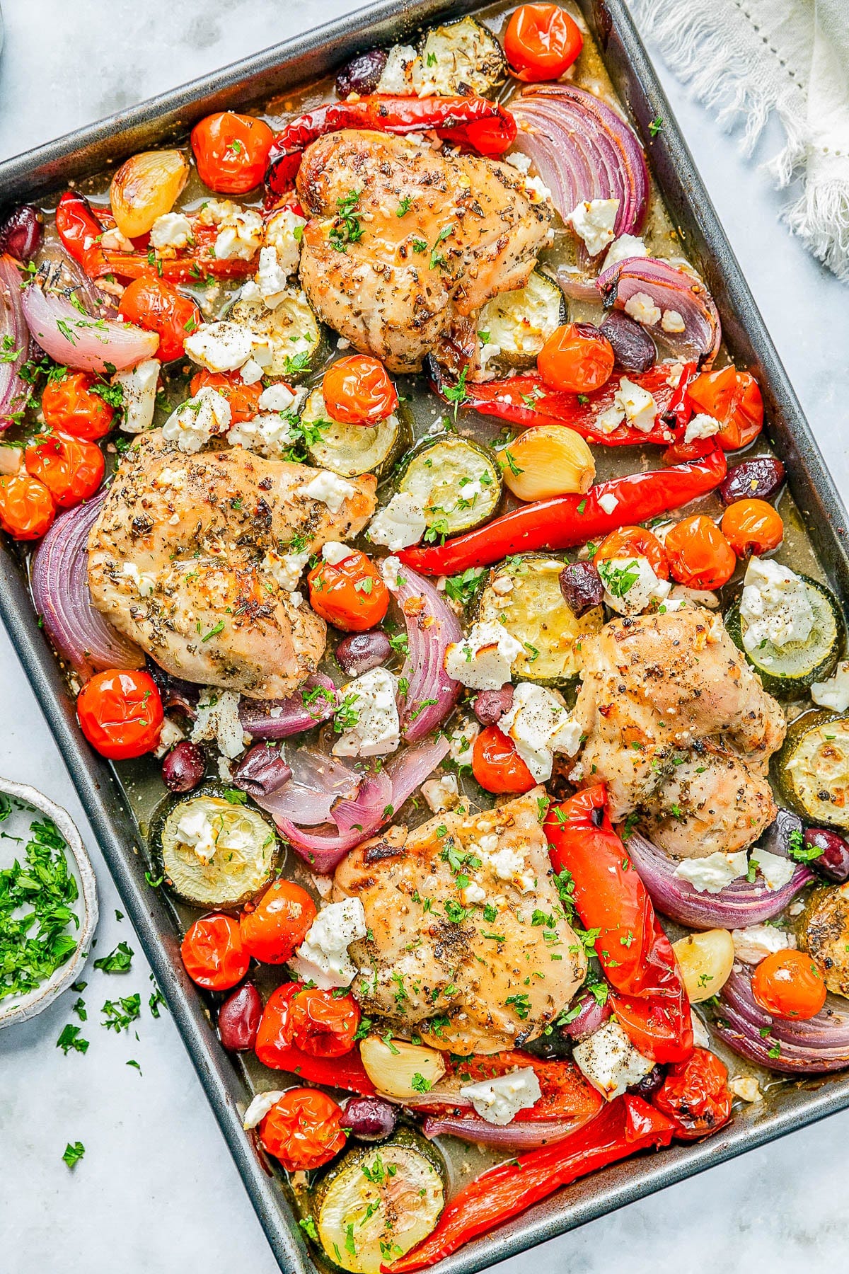 Baked chicken thighs with cherry tomatoes, zucchini, red onions, red bell peppers, olives, and crumbled feta on a sheet pan, garnished with chopped parsley. Baked chicken thighs with cherry tomatoes, zucchini, red onions, red bell peppers, olives, and crumbled feta on a sheet pan, garnished with chopped parsley.