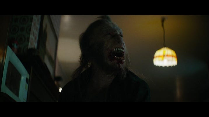 Wolf Man transformation scene — Practical effects shot of the werewolf mid-transformation, snarling with bloodied teeth inside a dimly lit farmhouse.