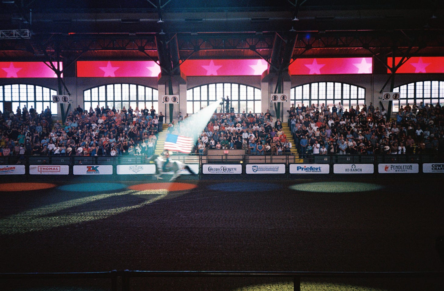Woman in a sparkly red shirt riding a brown horse around the rodeo arena of Cowtown Coliseum in Fort Worth, Texas. Under a single spotlight carrying a giant American flag. Photographed by Connor Fowler on 35mm film.