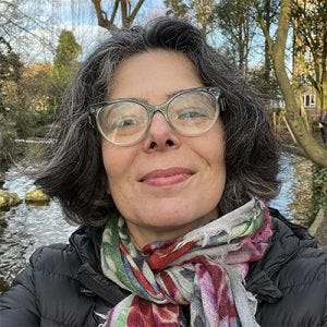 20260706_euniceostrensky Mulher de cabelos grisalhos curtos e volumosos usa óculos de grau de armação grossa e transparente. Ela veste uma jaqueta acolchoada preta e um cachecol vibrante e colorido com estampas florais. Ela está ao ar livre, em pé com árvores ao fundo em um plano médio e olha para a câmera com um sorriso suave.