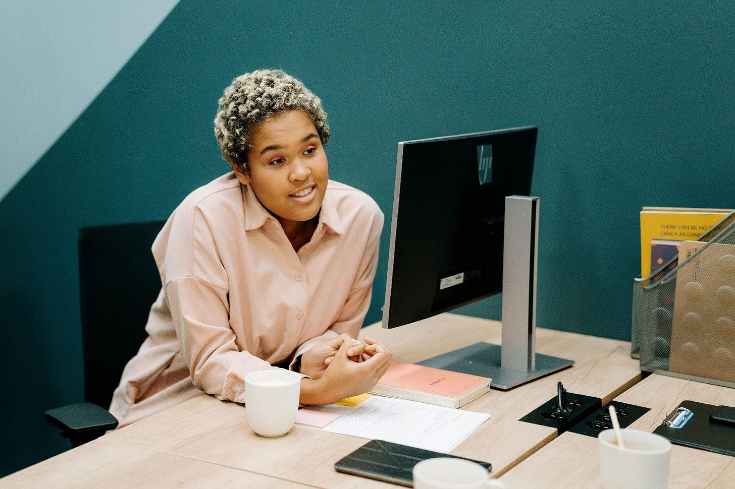dark skinned woman with short, curly, blond hair wears a long sleeve pink shirt and sits at a computer desk dark skinned woman with short, curly, blond hair wears a long sleeve pink shirt and sits at a computer desk