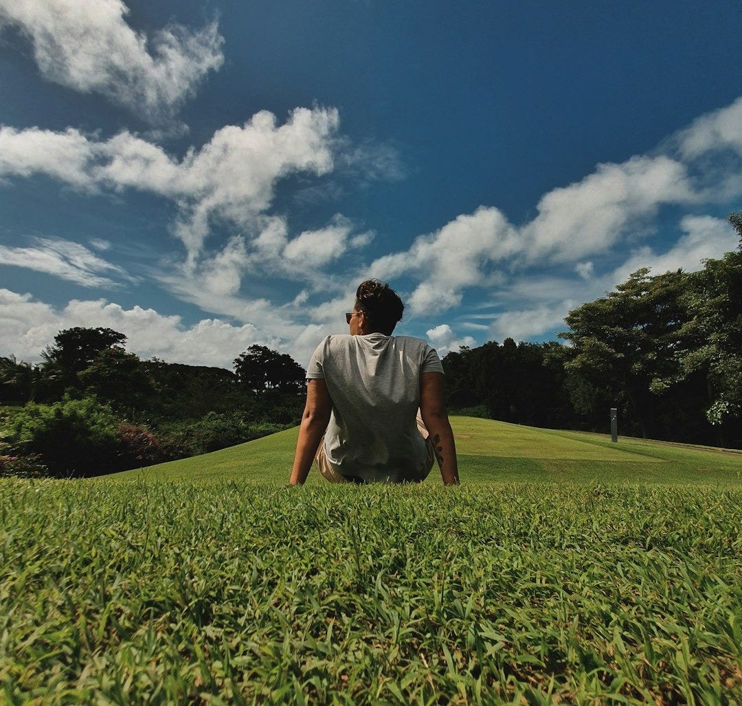 man in white t-shirt sitting on green grass field under blue and white cloudy sky