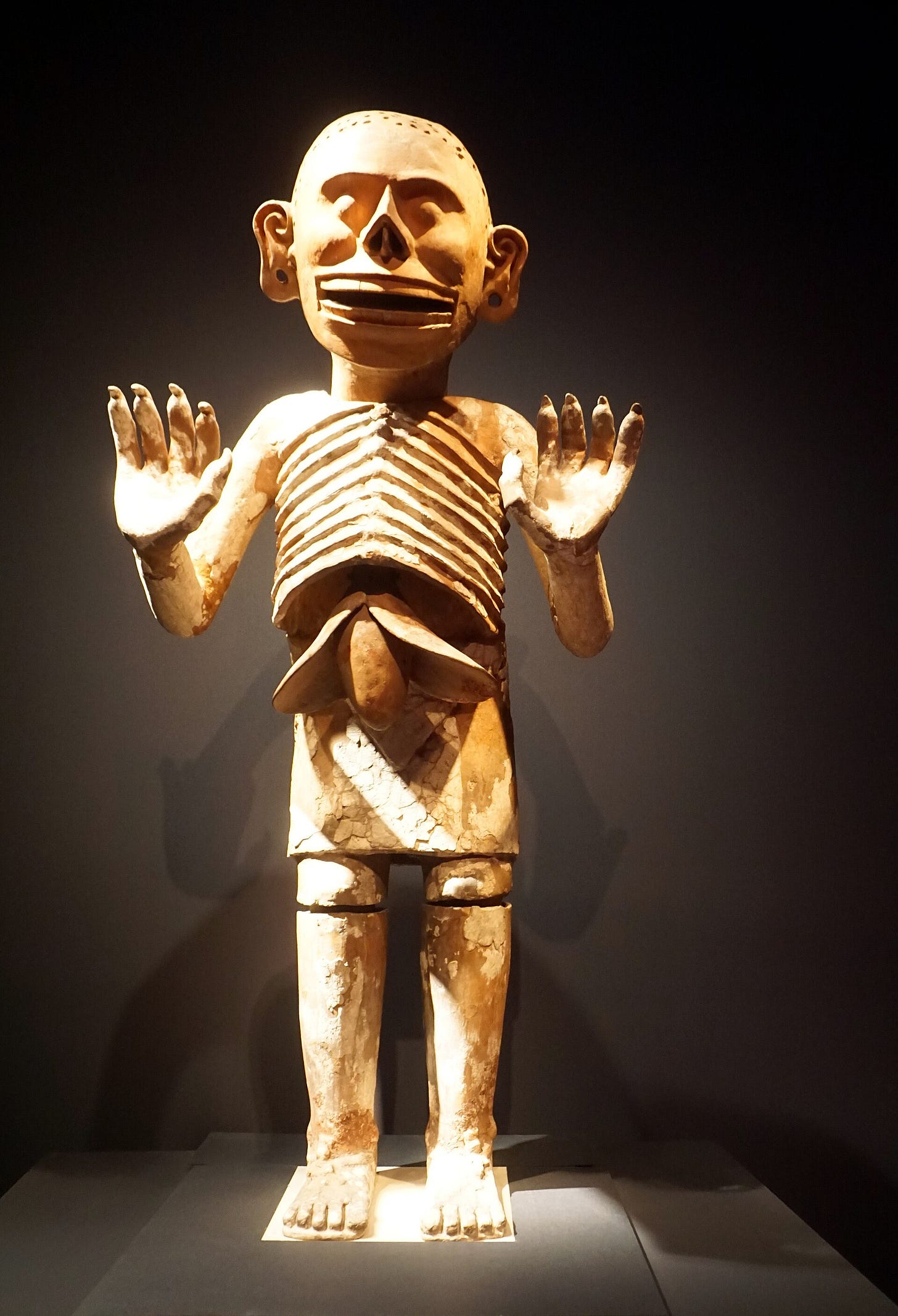 A large standing statue in beige clay, leaning forward with arms raised and clawed hands, its face a skull with bared teeth and a round shape hanging beneath the ribs. A large standing statue in beige clay, leaning forward with arms raised and clawed hands, its face a skull with bared teeth and a round shape hanging beneath the ribs.