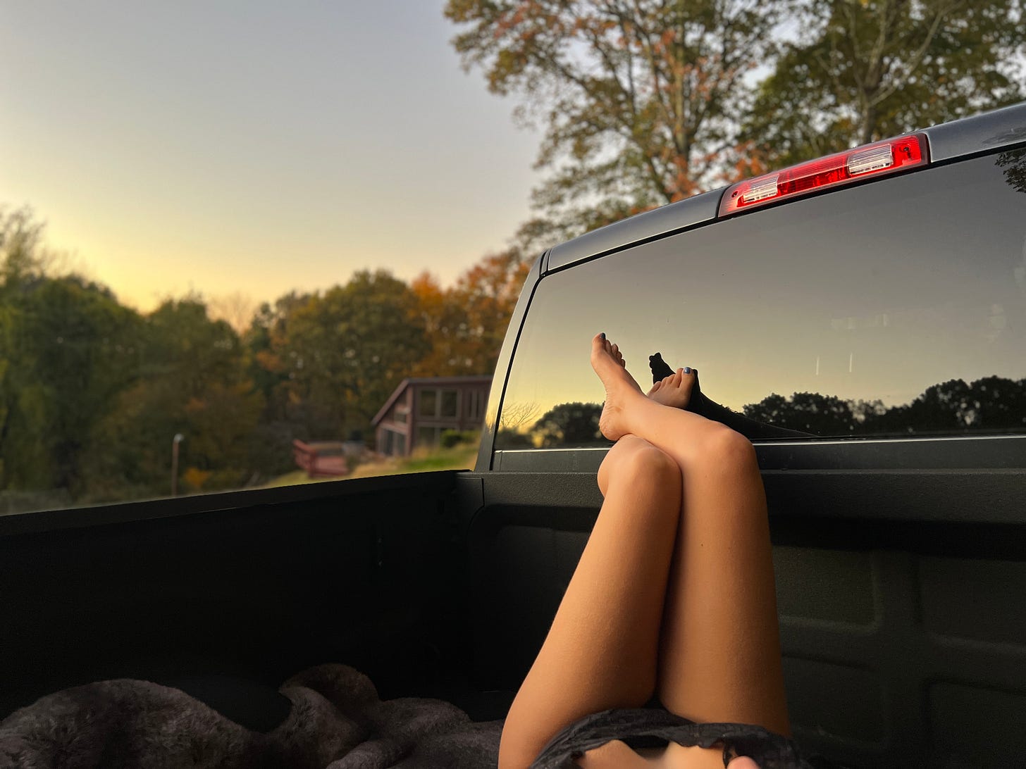 Torso in black lingerie lying in the back of a truck, legs propped along the edge, surrounded by trees, sunset, and a beautiful retreat center