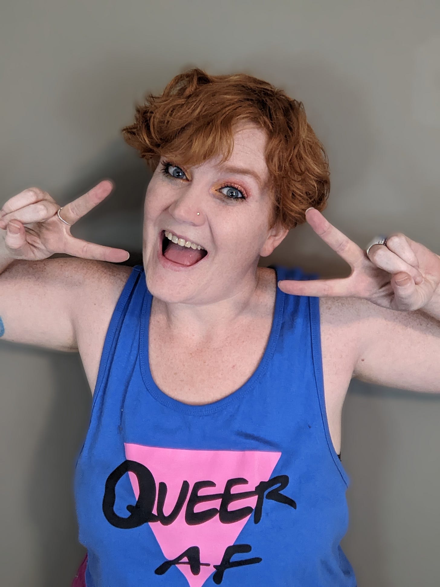 A picture of a non-binary person with curly red hair flashing peace signs and smiling at the camera. They are wearing a blue tank top with an inverted pink triangle that says "Queer AF."