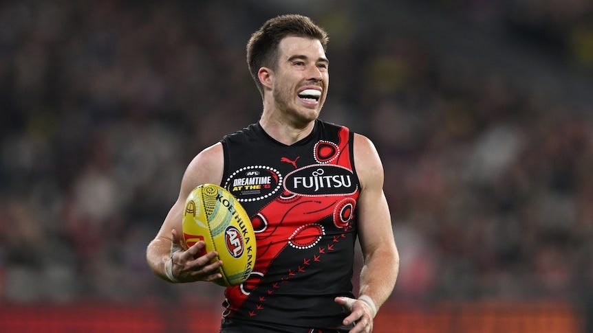 Zach Merrett of Essendon holding the AFL ball under one arm, looking to kick to a teammate Zach Merrett of Essendon holding the AFL ball under one arm, looking to kick to a teammate