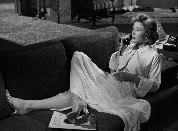 Gloria Grahame lounges on a mid-century couch, elegantly dressed in a robe and heels, talking on a rotary phone with a magazine at her feet. The scene oozes noir glamour.