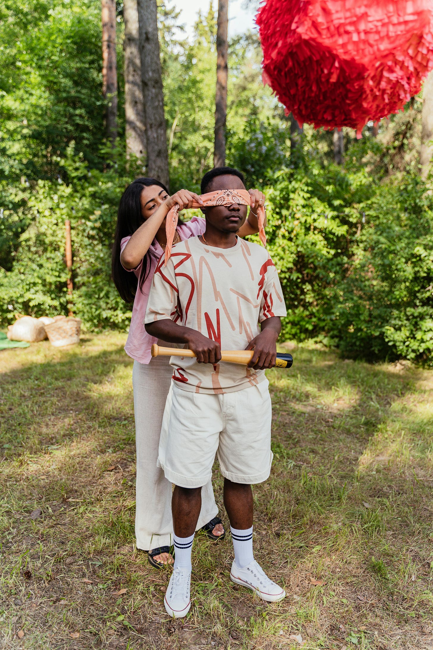 A man being blindfolded while holding a bat in a garden. There is a pinata in the foreground. A man being blindfolded while holding a bat in a garden. There is a pinata in the foreground.