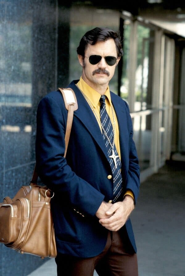 John Stockwell, with short, dark hair and a mustache, stands outside a marble building, wearing dark aviator glasses, a yellow shirt, striped tie and blue blazer, carrying a brown messenger bag. John Stockwell, with short, dark hair and a mustache, stands outside a marble building, wearing dark aviator glasses, a yellow shirt, striped tie and blue blazer, carrying a brown messenger bag.