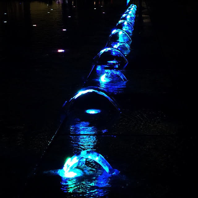 Night photo with lighted blue fountains that are close the ground, running in arch shapes and trickling