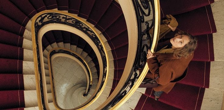 A red-carpeted spiral staircase with gilded wrought-iron railing, seen from above; a woman in a brown coat pauses partway down, looking up toward the camera. A red-carpeted spiral staircase with gilded wrought-iron railing, seen from above; a woman in a brown coat pauses partway down, looking up toward the camera.