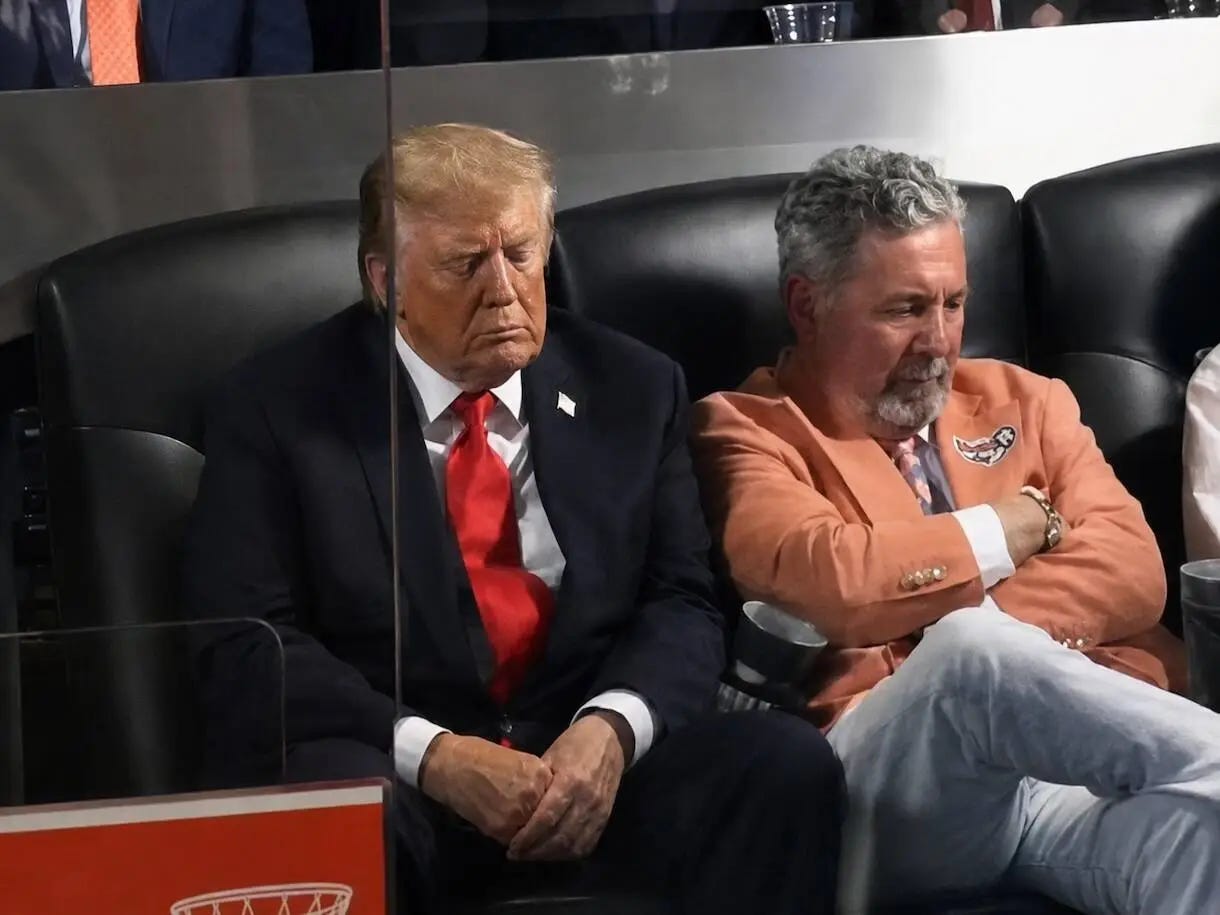 Two men sit side by side in black leather seats; the man on the left wears a dark suit with a red tie and has eyes closed in a thoughtful pose.