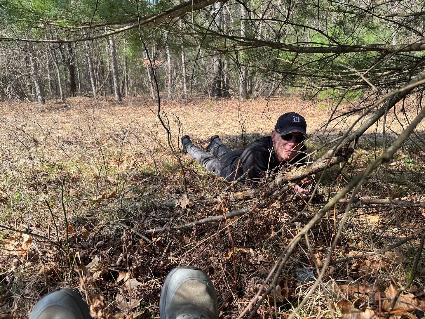 A view of the woods in midmorning sunshine, the author's feet visible and her intrepid life partner on his belly in the grass in front of her, just at the front edge of the pine tree she's sitting under. He's wearing a Detroit Tigers ballcap and smiling.
