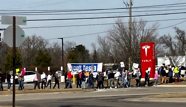 Protestors walk with picket signs in front of a Tesla dealership in Ann Arbor, Mich. The dealership is a long white building with the word Tesla. People with placards march in front of the building.