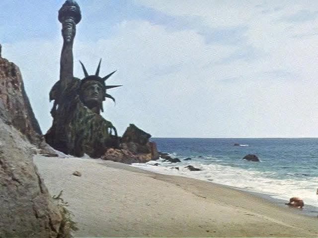 A large statue resembling the Statue of Liberty appears decayed and covered in green moss and vines with its torch arm extended upward and the face partially obscured by growth standing on a rocky cliffside overlooking a sandy beach and calm ocean waves under a partly cloudy sky a small brown dog walks on the sand in the foreground rocks and distant sea elements are visible
