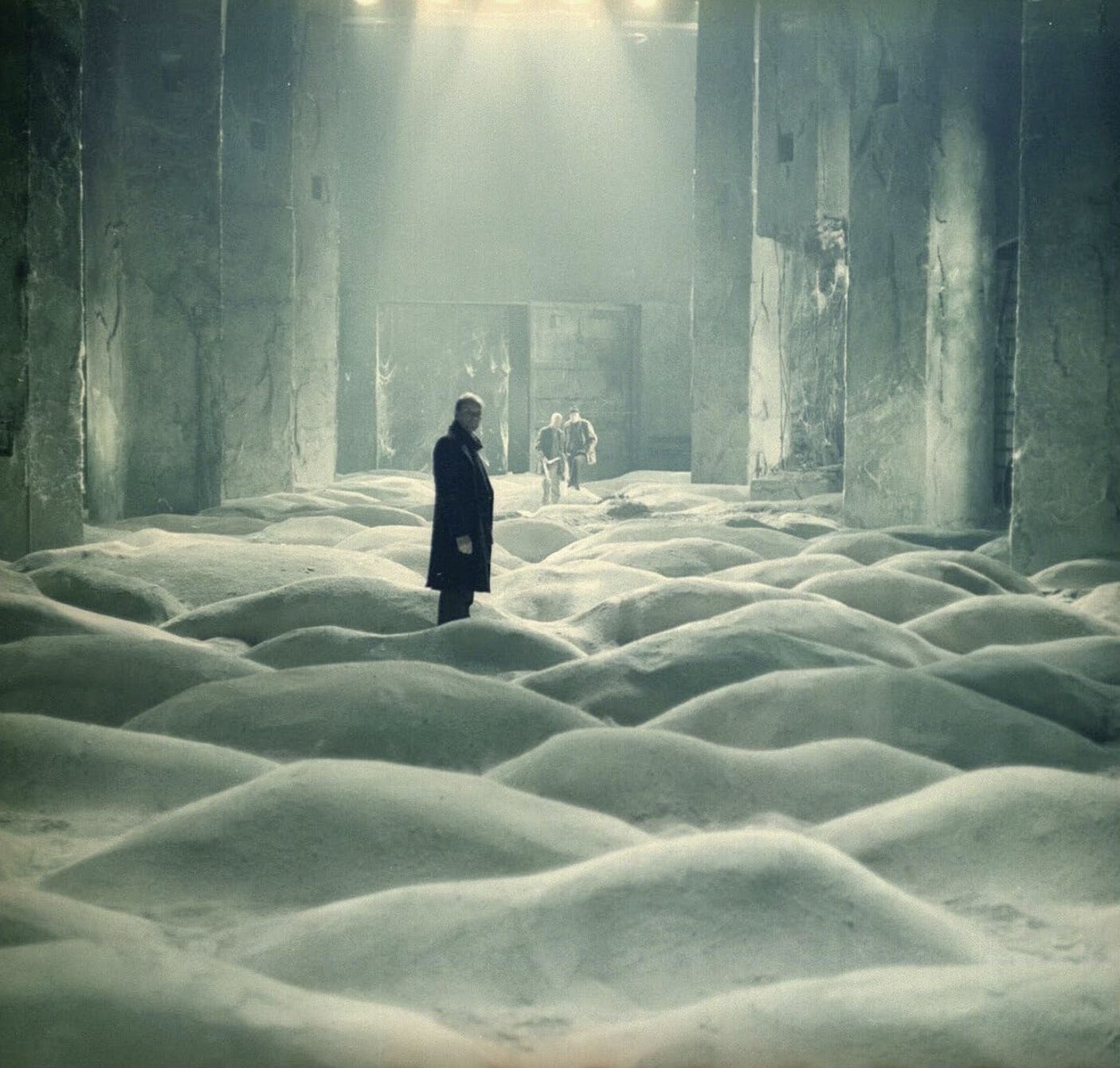 The Writer (Anatoly Solonitsin) standing in the middle of what appears to be an outcrop of sand dunes inside a cavernous room, with the Stalker (Aleksandr Kaidanovsky) and the Professor (Nikolai Grinko) watching him from a distance.