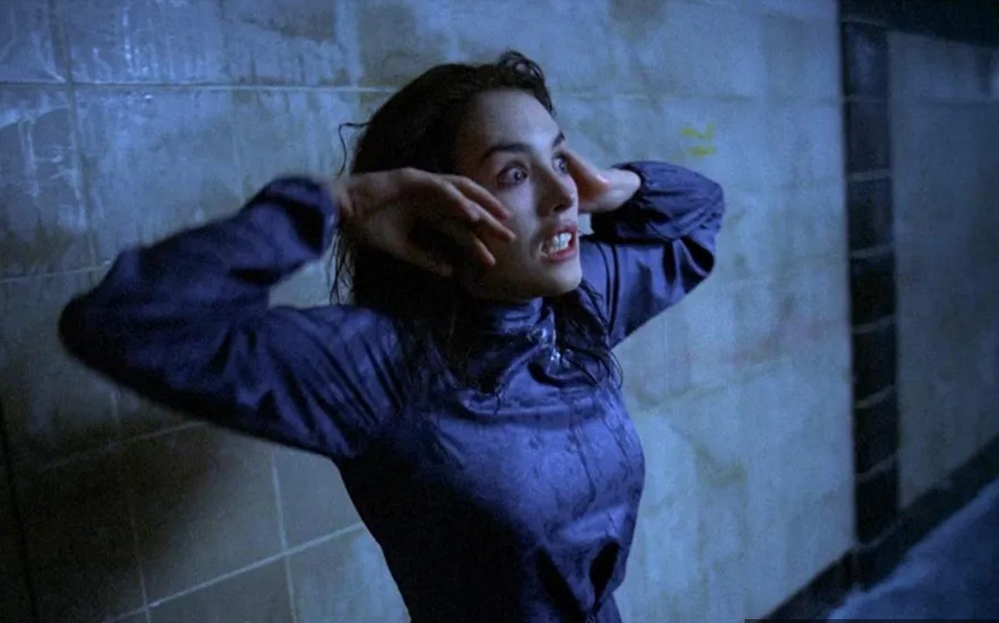 A woman in a blue dress holds her hands next to her face with a wide-eyed, crazed expression.