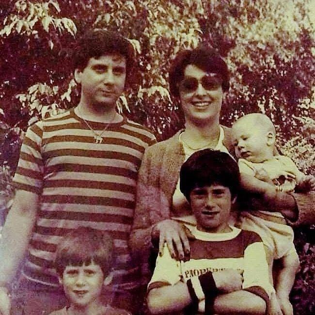 A vintage family photo showing a couple with their three young sons outdoors. The father stands beside the mother, who holds a baby, while two boys stand in front. The image conveys warmth, resilience, and the subtle space that defined their love.