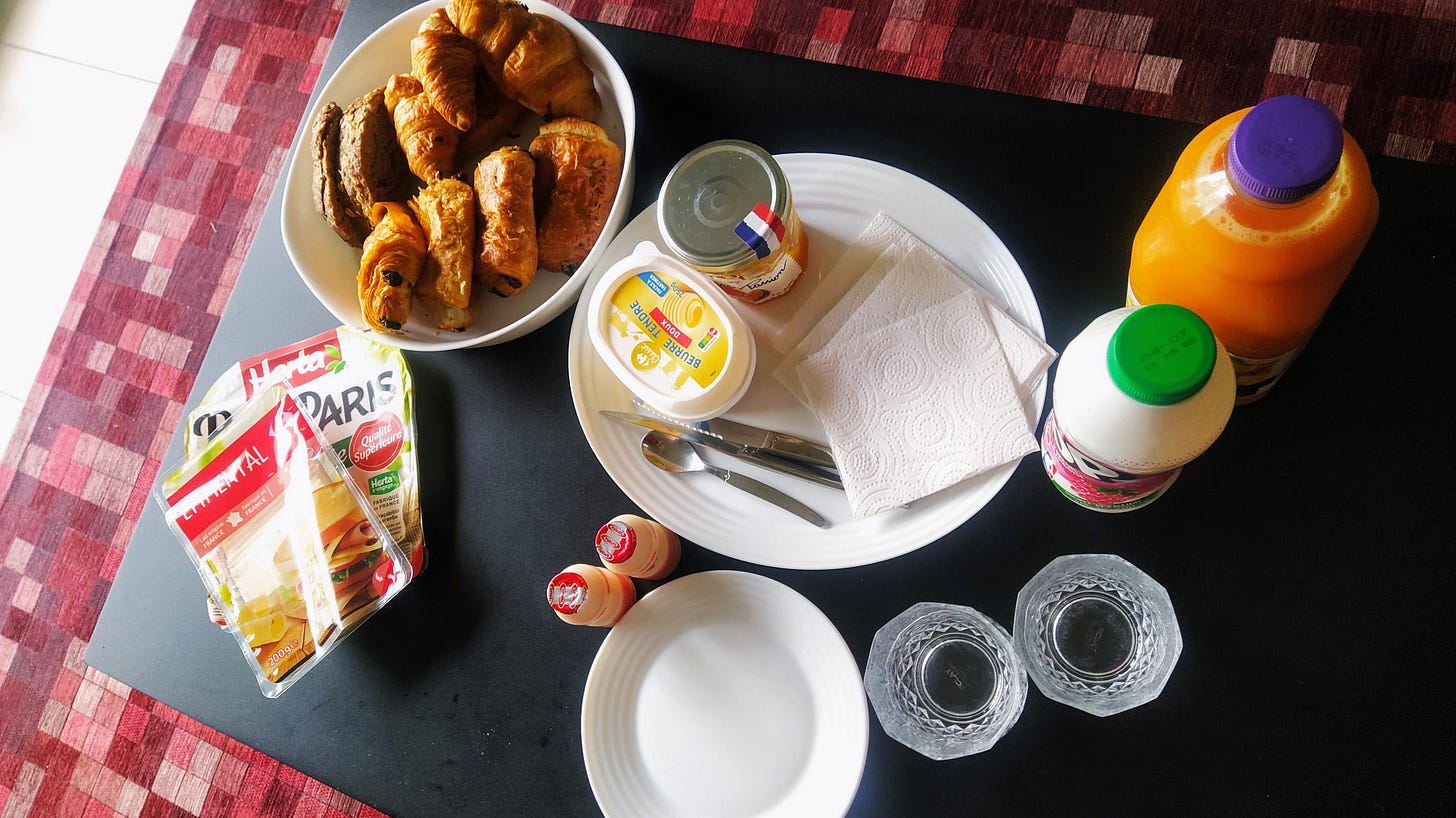 Looking from the top of a black coffee table. There’s a bowl filled with croissants and pain au chocolat, and packets of cheese and ham beside it. Next to it there’s butter, jam, cutlery and napkins on a white dish. To the side there’s two bottles, one of orange juice and another of yogurt. Two probiotic bottles, a smaller dish and two glasses complete the ensemble. Looking from the top of a black coffee table. There’s a bowl filled with croissants and pain au chocolat, and packets of cheese and ham beside it. Next to it there’s butter, jam, cutlery and napkins on a white dish. To the side there’s two bottles, one of orange juice and another of yogurt. Two probiotic bottles, a smaller dish and two glasses complete the ensemble.