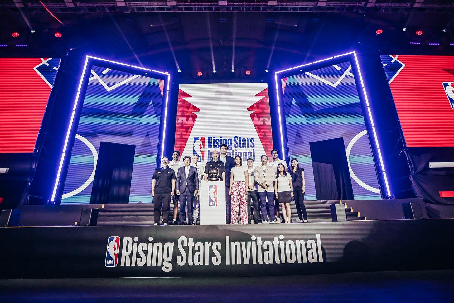  A wide, low-angle shot of a group of people, including NBA officials and players, posing on a grand stage at the NBA Rising Stars Invitational in Singapore. The group is gathered behind a white podium featuring the NBA logo and a basketball trophy. In the background, three massive LED screens display vibrant blue, red, and white graphics with the "Rising Stars Invitational" branding. The front of the stage is draped in a black banner with the tournament's title in bold white lettering. Bright stage lights and red spotlights create a professional, high-energy atmosphere for the event's opening ceremony.