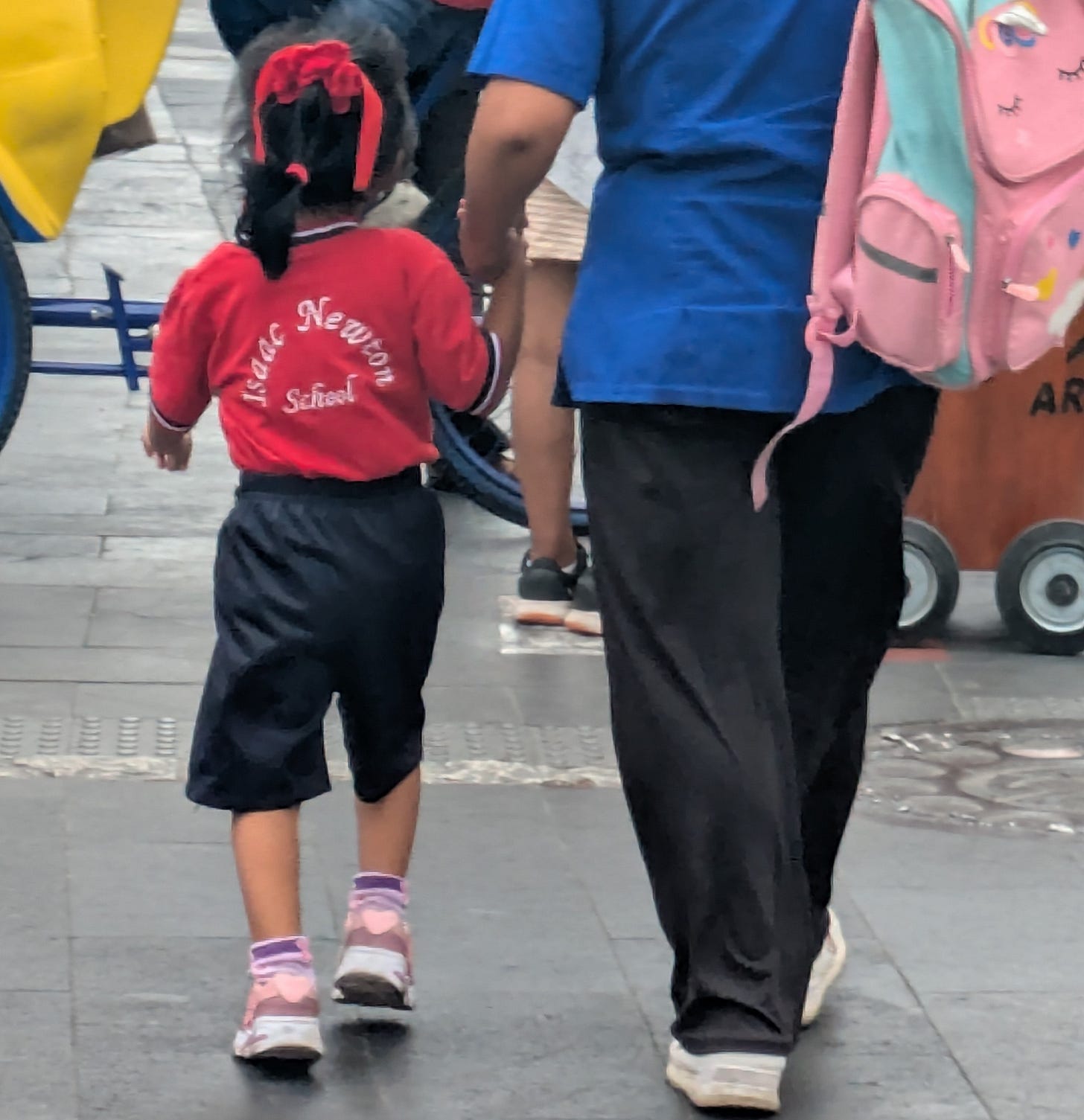 Photograph of a little girl in Lima, Peru wearing a red school shirt and dark shorts, and with a red ribbon in her hair, walks after school with her grandmother.