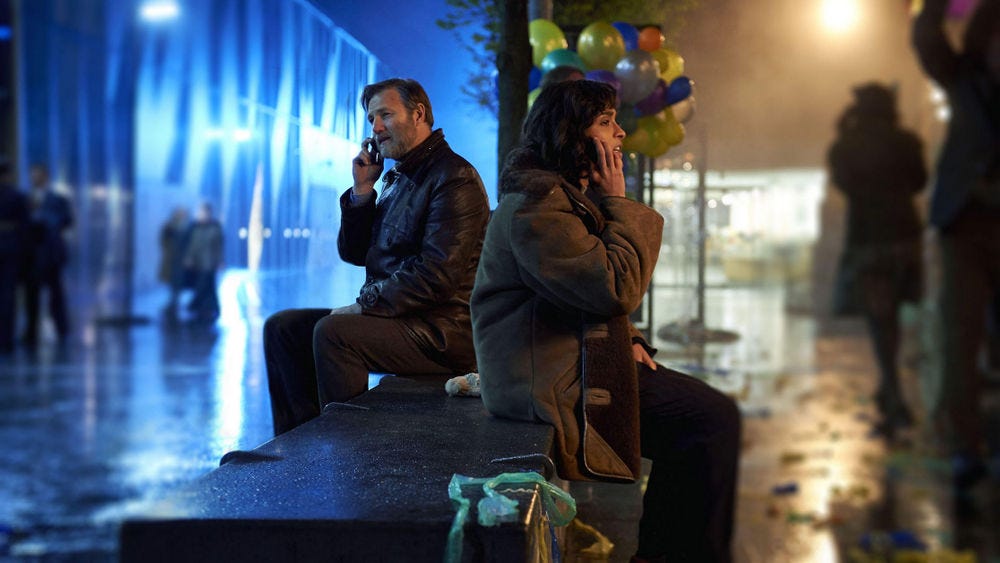 A scene from the BBC's adaptation of The City & The City, showing two detectives sat on opposite sides of a park bench, unable to talk to each other directly because they're in different cities, but able to talk on their mobile phones.