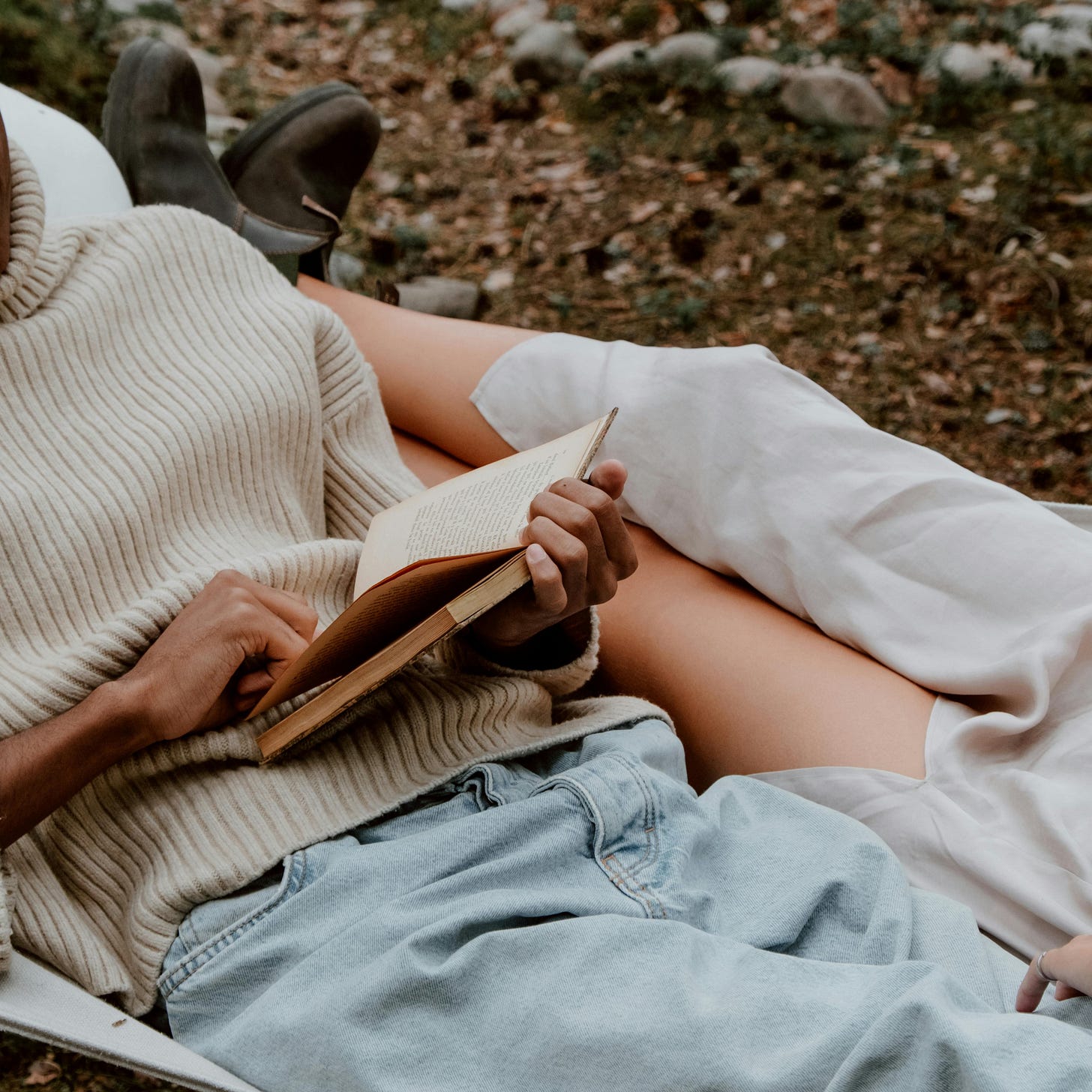 A person in a cosy sweater reads a book while lounging beside another person, in a serene outdoor setting.